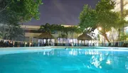 An illuminated swimming pool area in the evening, surrounded by tropical trees and lounging facilities, reflects the tranquil ambiance of a resort-like setting.