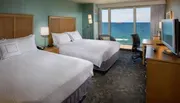 A hotel room with two beds features a large window offering a panoramic view of the ocean.
