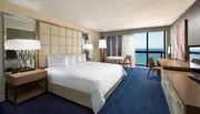The image shows a well-appointed hotel room with a large bed, modern furniture, and a balcony overlooking a blue ocean.