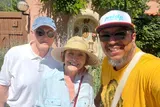 Three smiling people are posing together for a photo in a sunny outdoor setting with a decorative niche in the background.
