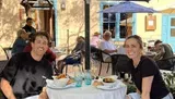 Two people are smiling at the camera while sitting at a table with food and drinks on a sunny outdoor patio at a restaurant.