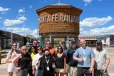 A group of smiling individuals is posing for a photo in front of a wooden water tank marked Santa Fe Railyard under a clear blue sky.