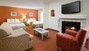 The image shows a well-furnished hotel room with twin beds, a seating area, a fireplace, and a wall-mounted flat-screen TV.