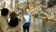 A couple is taking a selfie by a canal with a stone bridge in the background, while an older man stands in the foreground looking towards the camera.