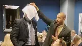 A man is playfully lifting a napkin over the head of another person, whose head is not visible, creating a humorous and candid moment in a social setting.