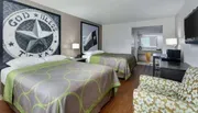 The image shows a hotel room with two beds featuring Texas-themed decor, with a God Bless Texas wall emblem, patterned bedspreads, and a seating area.