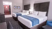 The image shows a neatly arranged hotel room with two double beds, featuring white linens with blue patterned runners, a small seating area, and artworks on the wall, creating a tidy and inviting space.