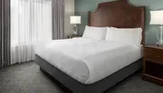 The image shows a neatly made bed with white linens in a tidy room with a classic decor style, featuring a large headboard, bedside tables, and lamps.