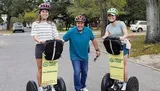 Three people are posing with Segways on a residential street, wearing helmets and smiling, with the signage indicating they are part of a tour group.