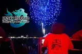 A person on a boat watches a vibrant blue firework display over the water at night, with a Frisky Mermaid Dolphin Tours & Boat Rentals logo in the upper left.