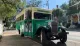 A vintage-style green and cream 'Hop On Hop Off' tour bus is parked on a sunny, tree-lined street with historic houses in the background.