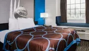 The image shows a well-kept hotel room with a bed that has a brown and blue patterned bedspread, a blue accent wall, and the words Pensacola Beach on a decorative pillow.