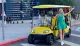 A smiling woman in a green dress is standing next to a yellow golf cart on a city street, gesturing with her arm outstretched.