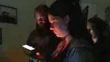 In a dimly lit room, a person is looking intently at their smartphone screen, illuminating their face, while another person appears in the background with a faint glow on their face.