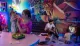 A family enjoys time together in a vibrantly colored room, where a young child and a woman play with an old-fashioned telephone, and another child and a man interact with a vintage television.