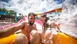 A man and a woman are joyfully riding down a water slide at a water park, with splashes of water around them.