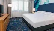 The image shows a modern hotel room with a neatly made bed, a television on a wooden cabinet, and a large abstract blue wall art above the headboard.