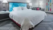 The image shows a neatly made king-size bed in a modern hotel room with contemporary decor and artwork.