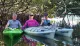 Three people are smiling while sitting on two kayaks among mangroves, with calm waters reflecting the scene.