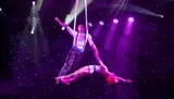 A performer gracefully hangs in mid-air held by her partner against a backdrop of theatrical lighting and a starry background, creating an illusion of flying in a dramatic stage performance.