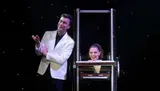A magician gestures towards his assistant, whose body appears to be magically severed at the waist by an illusion box on stage.