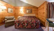 The image shows a cozy bedroom with wood-paneled walls, a colorful quilt on the bed, plaid upholstered furnishings, and framed artwork, creating a rustic ambiance.