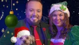 A couple dressed in festive holiday attire poses with a small dog wearing a Santa hat, set against a starry night sky with a hanging green ornament.