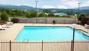 This image shows an outdoor swimming pool enclosed by a fence with a few lounge chairs, a single person sitting at the far end, and hilly terrain in the background under a clear sky.