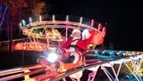 A person dressed as Santa Claus is joyfully waving while riding a small roller coaster at nighttime, illuminated by colorful lights.