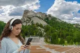 A young woman wearing headphones is focused on her phone in the foreground, with the iconic Mount Rushmore National Memorial visible in the background.
