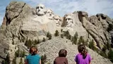 Three individuals are observed from behind gazing upon the iconic Mount Rushmore National Memorial, which features the monumental carvings of the faces of four U.S. presidents.