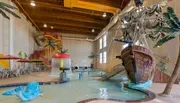 The image features an indoor water park with a pirate ship-themed play structure, a waterslide, and several seating areas under umbrellas.