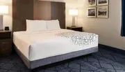 The image shows a neatly made bed with white bedding and a geometric pattern at the foot, in a clean and modern hotel room with pictures on the wall and a side table with a lamp.