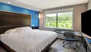 The image shows a modern hotel room with a large bed, a blue accent wall, a work desk with a chair, and a window providing a view of green hills.