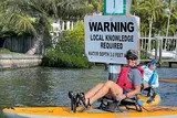 Two people in a kayak are passing by a warning sign indicating that local knowledge is required and specifying a water depth of 3.0 feet.
