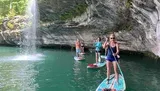 A group of people are paddleboarding near a waterfall and a rocky cliff.