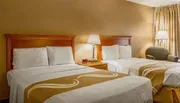The image shows a neatly arranged hotel room with two double beds, warm lighting, and simple decor.