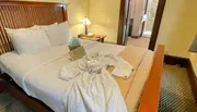 The image shows a neatly made hotel room bed with a white bathrobe laid out on it, a gift bag on the side, and a glimpse of an en-suite bathroom and entryway in the background.