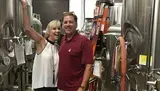 A smiling man and woman pose in front of stainless steel brewing equipment, suggesting a setting in a brewery.