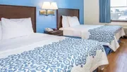 The image shows a neatly arranged hotel room with two twin beds covered with blue and white patterned bedspreads, a nightstand with a phone, and a window that allows natural light into the room.
