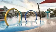 An empty water playground with colorful arches spraying water and a mushroom-shaped water feature, with loungers and buildings in the background, captured during the daytime.