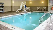 The image shows an indoor swimming pool with a mechanical lift device to assist individuals with disabilities in entering the water, surrounded by pool deck chairs and towels stacked on a shelf.