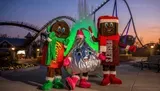 Three people in festive candy-themed costumes stand in front of a roller coaster at dusk.
