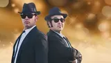 Two men dressed in black suits, hats, and sunglasses pose back-to-back with a blurred gold background.