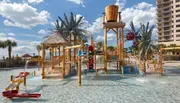 The image shows a colorful children's water playground with various water features, slides, and playful structures, located beside a beachfront high-rise building.