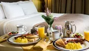 A sumptuous breakfast tray with scrambled eggs, bacon, toast, freshly sliced fruits, jam, orange juice, and coffee is elegantly presented on a bed with plush pillows and a warm ambiance.