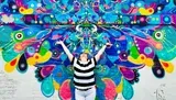 A person with arms raised stands in front of a vibrant, colorful mural with abstract patterns and eye motifs.