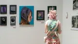 A person with pink hair and a whimsical outfit observes artwork displayed on a gallery wall.