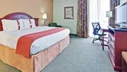 The image shows a tidily kept hotel room with a large bed, a desk with a chair, a television, and colorful curtains.