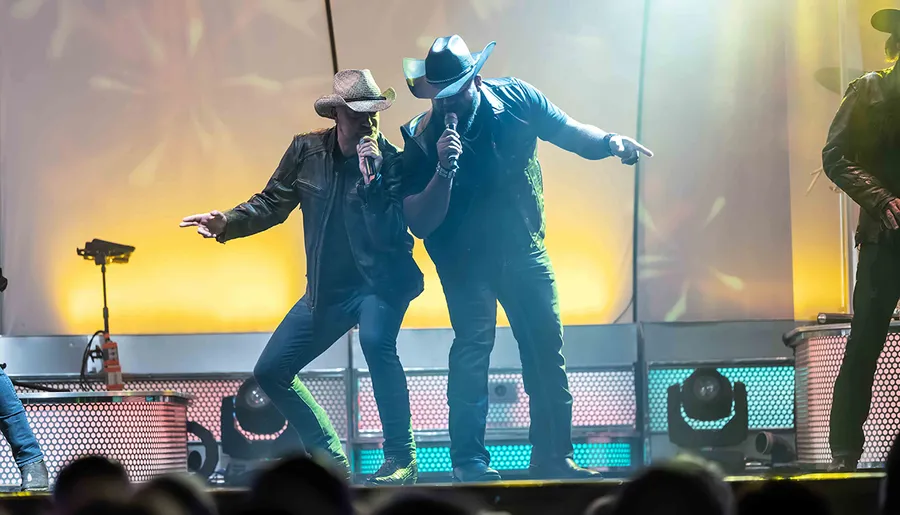 Two musicians in cowboy hats are energetically performing on stage with microphones in hand.